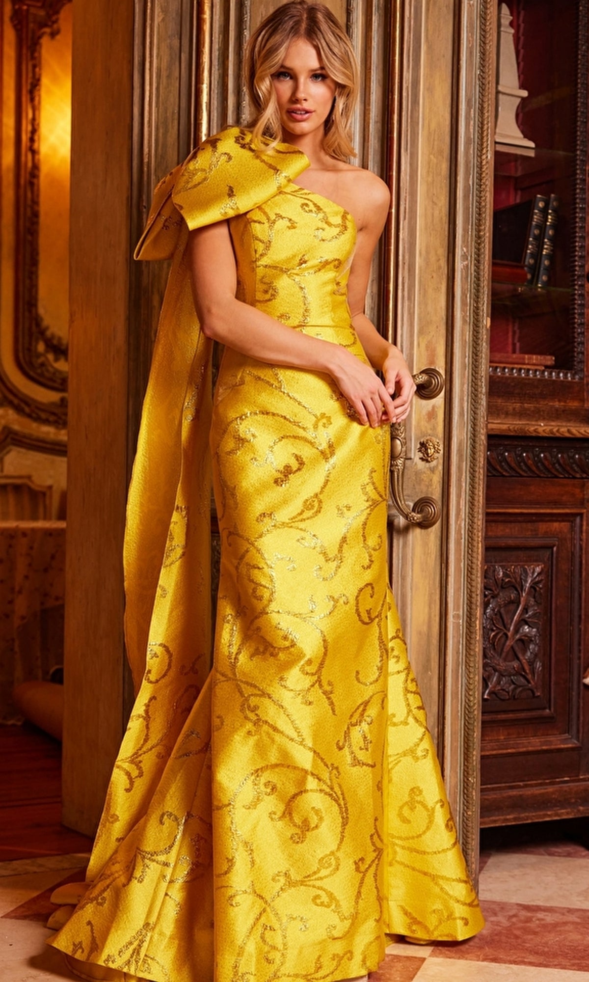 Woman wearing the Jovani One-Shoulder-Bow Long Formal Dress 23742 in yellow with an ornate pattern and large bow detail, standing indoors by a decorative door.